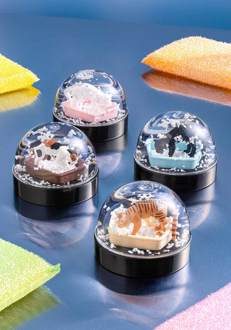 Four cat litter snow globes with cute cat figures inside and swirling white flakes on a shiny blue surface
