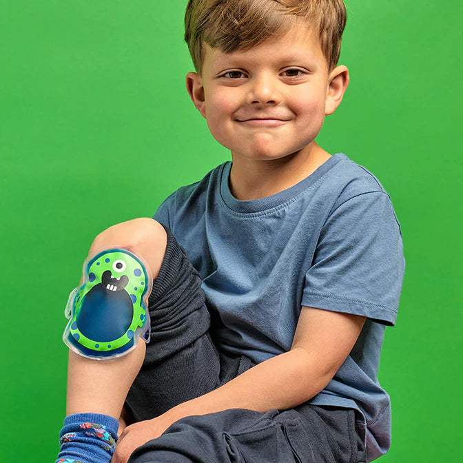 Young boy sitting with a colorful Oopsie Monster hot and cold pack on his knee against a green background