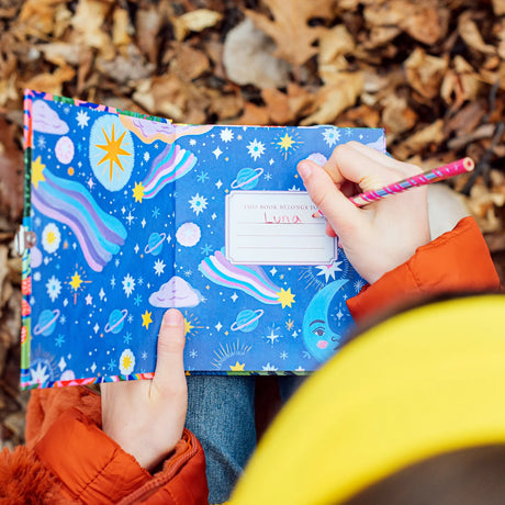 Person writing in a colorful foil-stamped journal with stars, planets, and whimsical space illustrations inside the cover