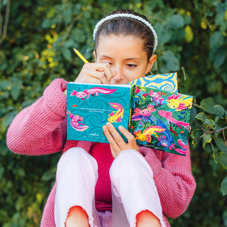 Young girl writing in colorful foil stamped Axolotl Diary journal with illustrated axolotls and lock in nature setting