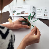 Person assembling a DIY paper banana plant from a kit with printed instructions on a white surface