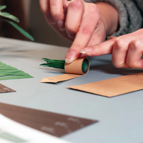 Hands carefully rolling paper strips to create DIY paper plants on a crafting table with green and brown sheets nearby
