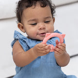 Baby in blue dress holding and playing with a pink pig-shaped teething rattle toy designed for infants.