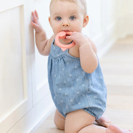 Baby in blue romper chewing on a peach pig-shaped rattle teether while standing near a white cabinet