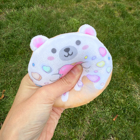 Hand squeezing a soft plush toy shaped like a donut with a cute bear face and colorful sprinkles outdoors on grass.