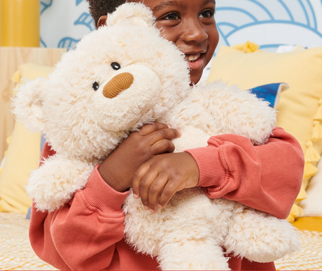 Child hugging a cream-colored Teddy Bear Bubbles plush with a corduroy nose, smiling happily indoors.