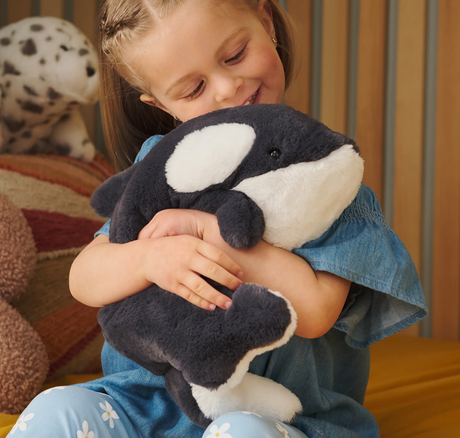 Child smiling and hugging a soft, black and white plush toy Snuffles Orca Flynn sitting upright.