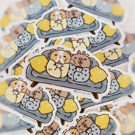 Bubu & Moonch Blanket Burritos Sticker showing a cozy guinea pig and capybara wrapped in blankets on a couch.