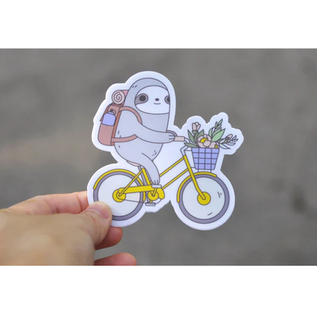 Hand holding a water-resistant Biking Sloth Sticker showing a sloth riding a yellow bike with flowers in the basket.