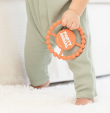 Baby in sage green textured romper holding orange beaded teething ring with white "PARTY ANIMAL" text on center disc