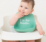 Smiling baby wearing a teal silicone bib with white text sitting in a white high chair against light background