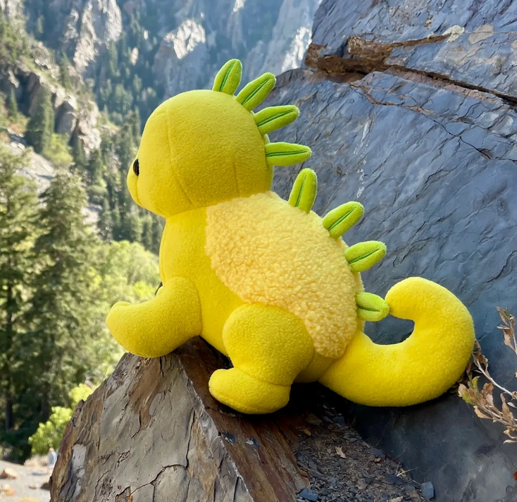 Yellow plush dinosaur with green fabric spikes and textured belly sitting on rocky ledge outdoors