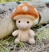 Small plush mushroom character with an orange cap and white spots sitting on grass beside a birch log