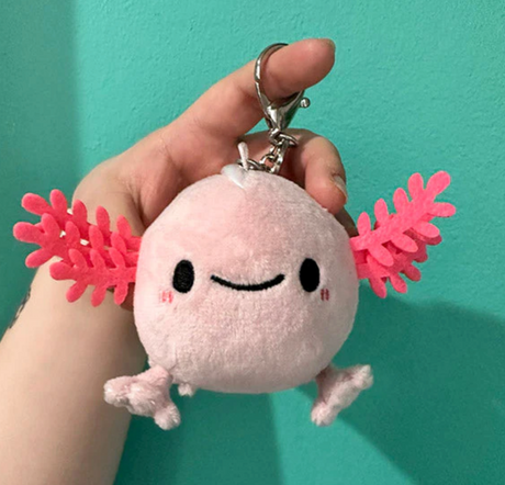 Hand holding a pink plush keychain shaped like a smiling axolotl with "Baby Axolotl Micro" product title.