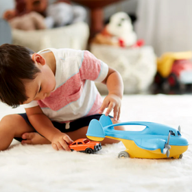 Child playing with a blue and yellow Cargo Plane toy and an orange car on a soft carpet indoors.
