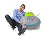 Man sitting on floor smiling next to large plush toy UFO Massive with bright green top and gray body