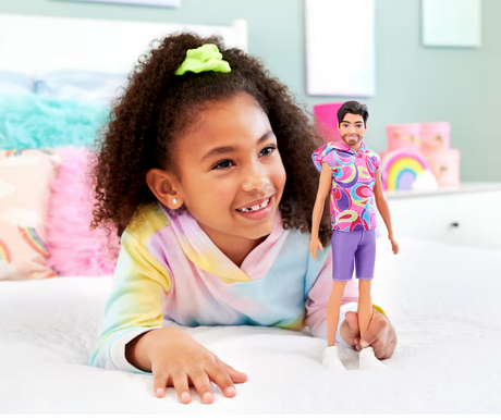 Girl playing with Barbie Fashionista Ken doll wearing colorful hoodie and purple shorts in bright room.