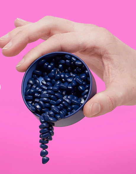 Hand holding a tin pouring indigo Speks. Matte. Mini Crags. magnetic putty pieces against a pink background.