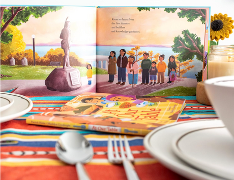 At Our Table children's book open to colorful page with diverse group honoring farmers, on a vibrant table setting.