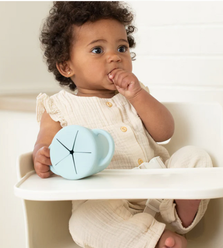 Baby holding a blue Happy Snacker | Cheerio Mate silicone snack cup with easy-grip handles in a high chair.