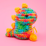 Woobles Crochet Kit Fred the Rainbow Dinosaur colorful crocheted dinosaur holding yarn and needle on pink background