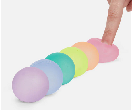 A finger pressing the pink gel ball from Speks. Gump Mini 6 set of six colorful mini stress balls.