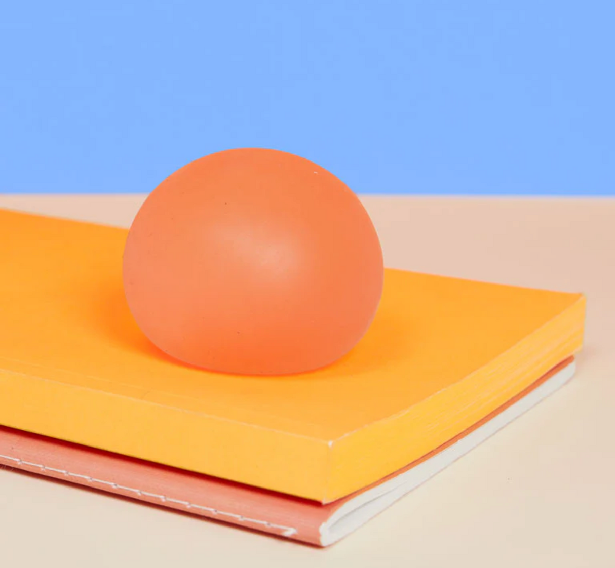 Speks. Gump Reef soft orange stress ball with gooey gel filling resting on stacked notebooks against blue background.