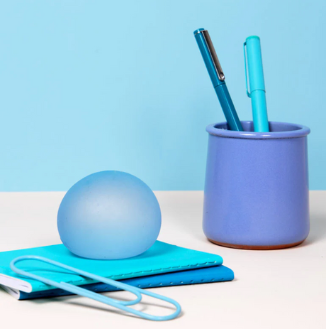 Speks. Gump Dew stress ball on a notebook beside a pen holder with two blue pens on a desk.