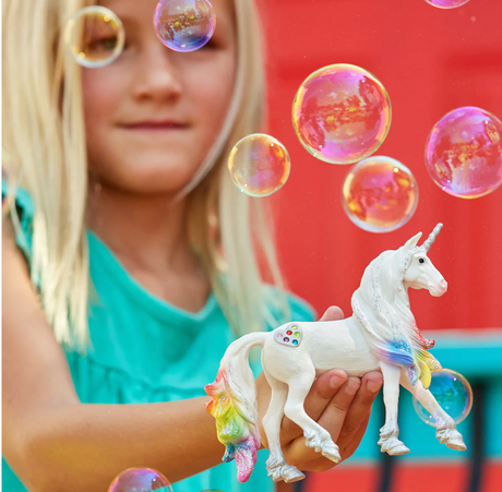 Child holding a white unicorn toy with rainbow mane and tail surrounded by bubbles, Bayala | Rainbow Love Unicorn Stallion