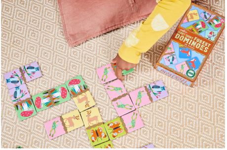 Child playing with Giant Shiny Forest Dominoes featuring colorful forest creatures on a woven rug beside the game box.