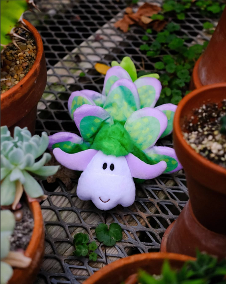 Leaf Sheep Emily plush toy with green and purple leaf-like body, smiling face, placed among potted plants outdoors.