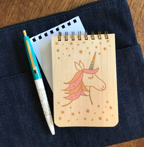 Mini Wood Notebook | Magical Unicorn with spiral binding and unicorn design, paired with a pen on dark fabric background.