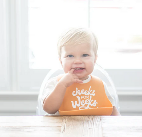Baby wearing an orange Wonder Bib | Cheeks For Weeks with catch pocket sitting at a wooden table.