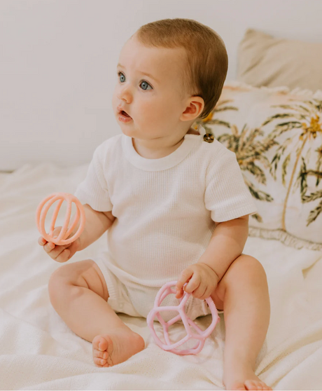 Baby sitting on bed holding Sensory Ball 2 Pack Bubblegum and Peach teether toys in each hand.