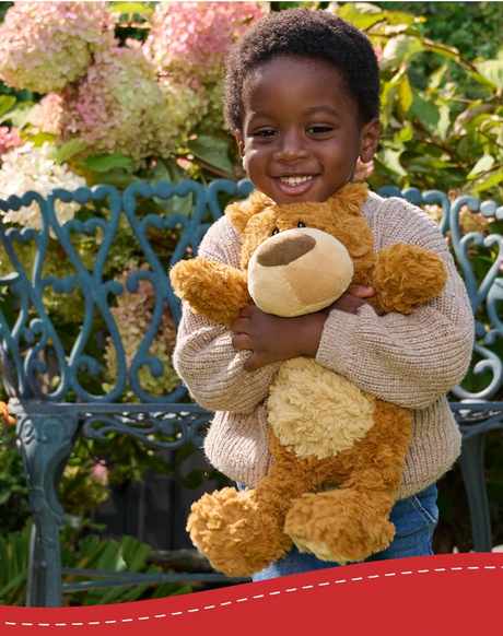 Child smiling and hugging a plush Teddy Bear Bexley with a tan tummy sitting outdoors on a sunny day.
