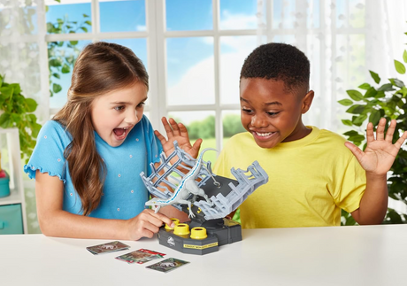 Two children excitedly playing Jurassic World Cage Breaker tabletop game with Velociraptor cage and action cards.