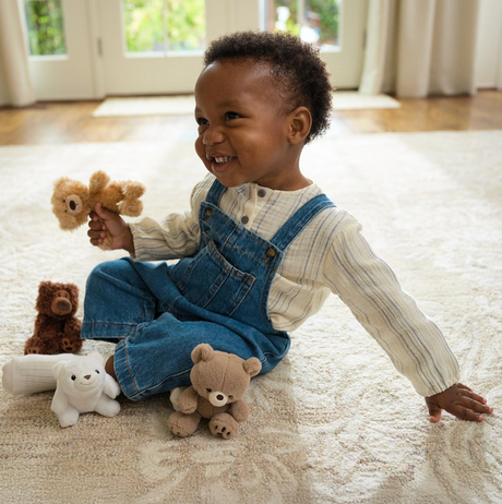 A smiling toddler plays with assorted Forever Friends Mini plush bears in a bright, cozy room.