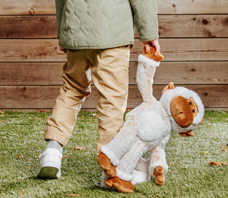 Child walking outdoors holding Monkey Tilly plush toy with soft coffee-and-cream fur and floppy limbs.