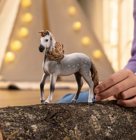 Andalusian Stallion toy model with gray coat and white spots, thick curly mane, posed on a log with hand nearby.