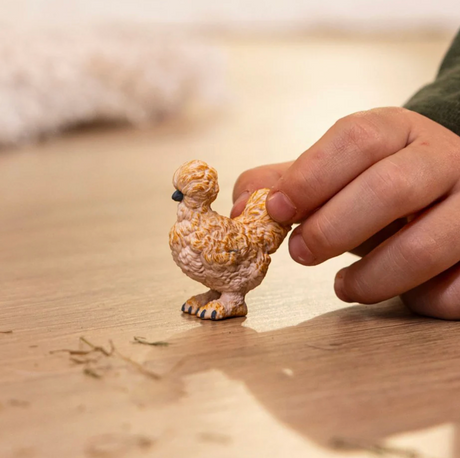 Child's hand holding a small Silkie Chicken toy with fluffy feathers and black beak on a wooden surface.