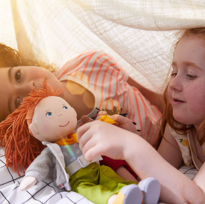 Soft Doll | Soley with red hair, cozy jacket, and green pants held by two children playing indoors.