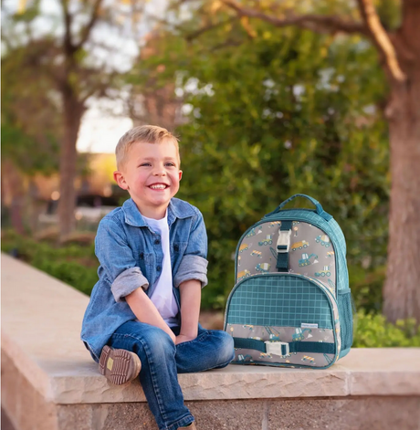 Happy young boy sitting outdoors next to a dinosaur print kids backpack with multiple compartments and straps