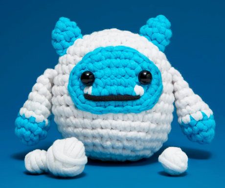 Chunky crocheted blue and white yeti with a toothy smile sitting on a blue background next to yarn balls