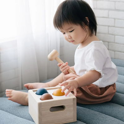 Toddler playing with a wooden pounding toy while sitting on a soft gray cushion by the window light
