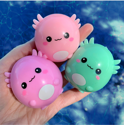 Hand holding three colorful squishy axolotl toys in pink, purple, and green with smiling faces by water background