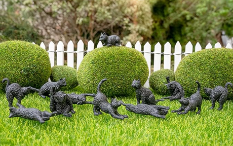 Twelve miniature black cat figurines in various playful poses arranged on grass with a white picket fence background
