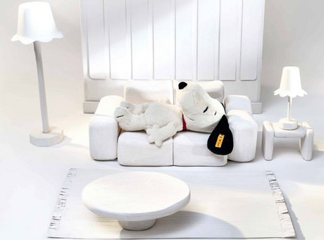 Soft plush Snoopy lying on a white couch in a minimalist all-white living room setting with lamps and table.