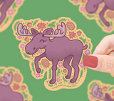 Hand holding a cute purple moose sticker surrounded by autumn leaves, pinecones, and flowers on green background