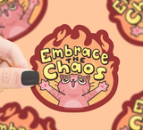 Hand holding a colorful 3-inch cat sticker with the phrase Embrace the Chaos and flames in the background