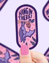 Hand holding a purple bat sticker hanging upside down from a branch with the words Hang in There on pink background